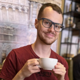 Kieran, a white man with short brown hair and dark blue glasses, holds a cup of coffee and smiles.